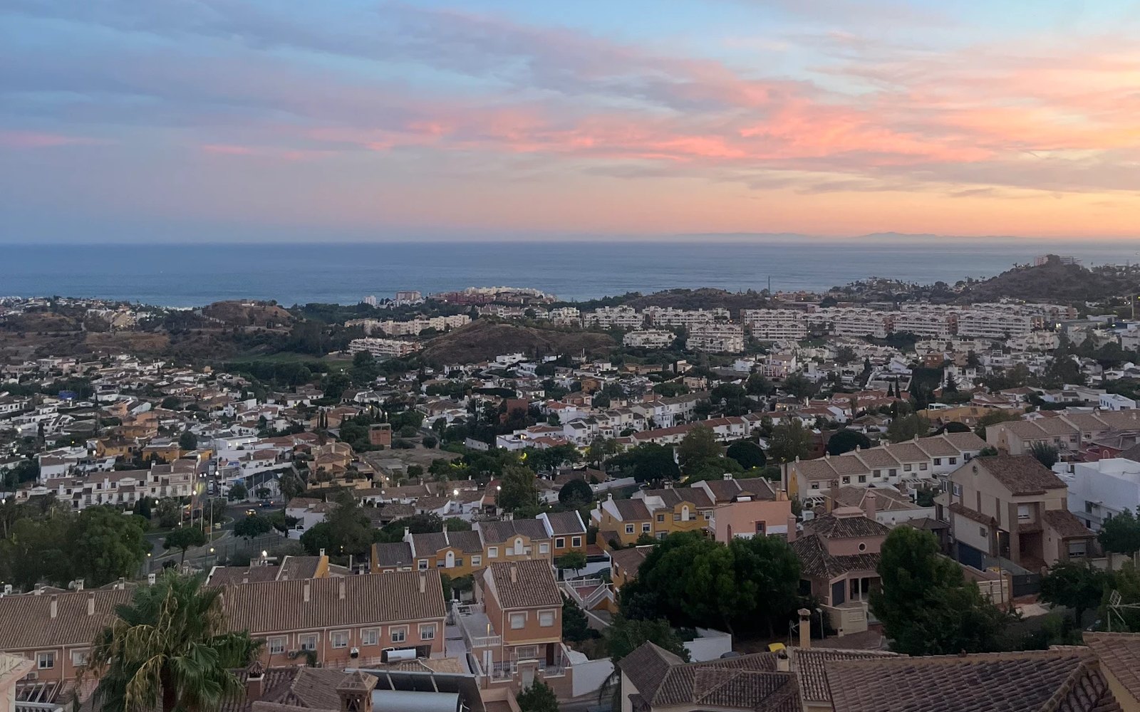 Vista al atardecer desde una urbanización en las alturas de la Costa del Sol, con tejados, el mar al fondo y la luna en un cielo rosado